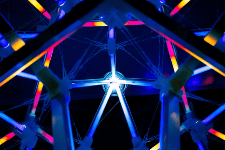 a large-scale geometric framework structure, as seen from the inside. The structure is lit in blue, pink, and yellow lights