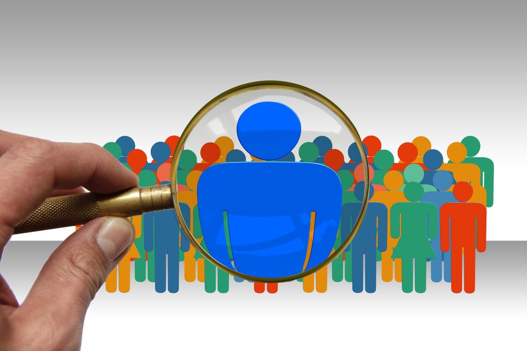 Icon-style large group of people. One person in middle highlighted by a magnifying glass.