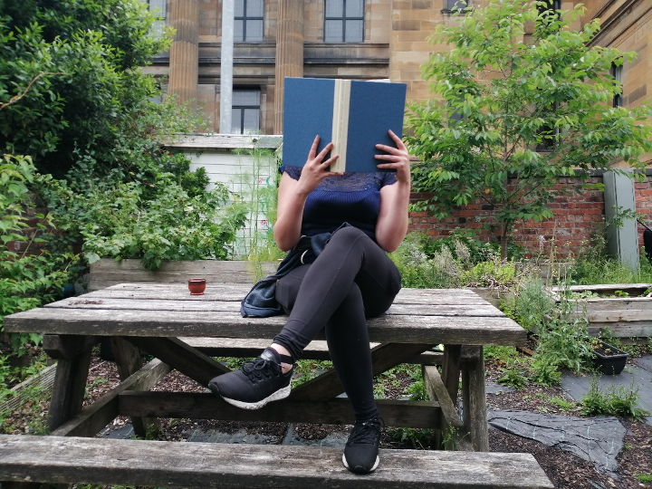 A person sits on a wooden table and bench in a garden, holding a large open hardback book in front of their face. 