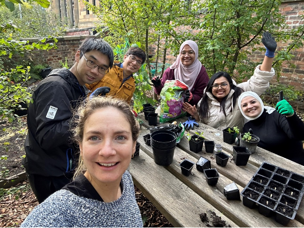 6 PGRs including Paola smile for the camera and wave. They are sitting around a picnic table, potting up small seedlings