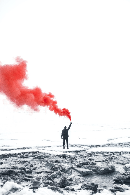 A figure stands at a distance, holding a red flare in an otherwise black and white snowy landscape.
