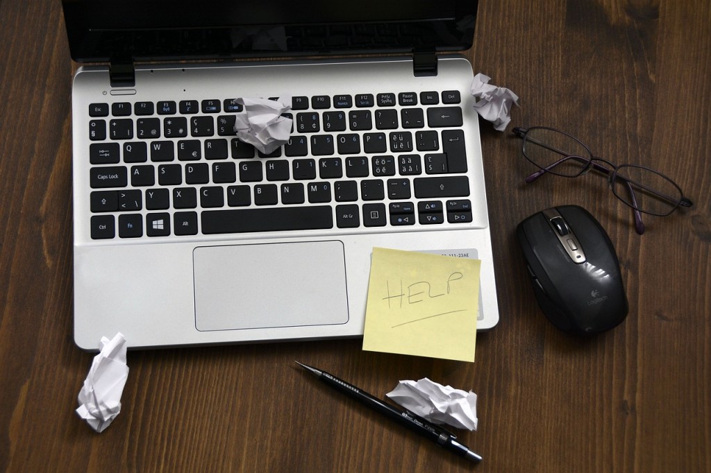 Image shows a laptop with a number of pieces of scunched up paper strewn around, and a Post-It note with the word 'HELP' written on it.
Image credit: anncapictures/pixabay.