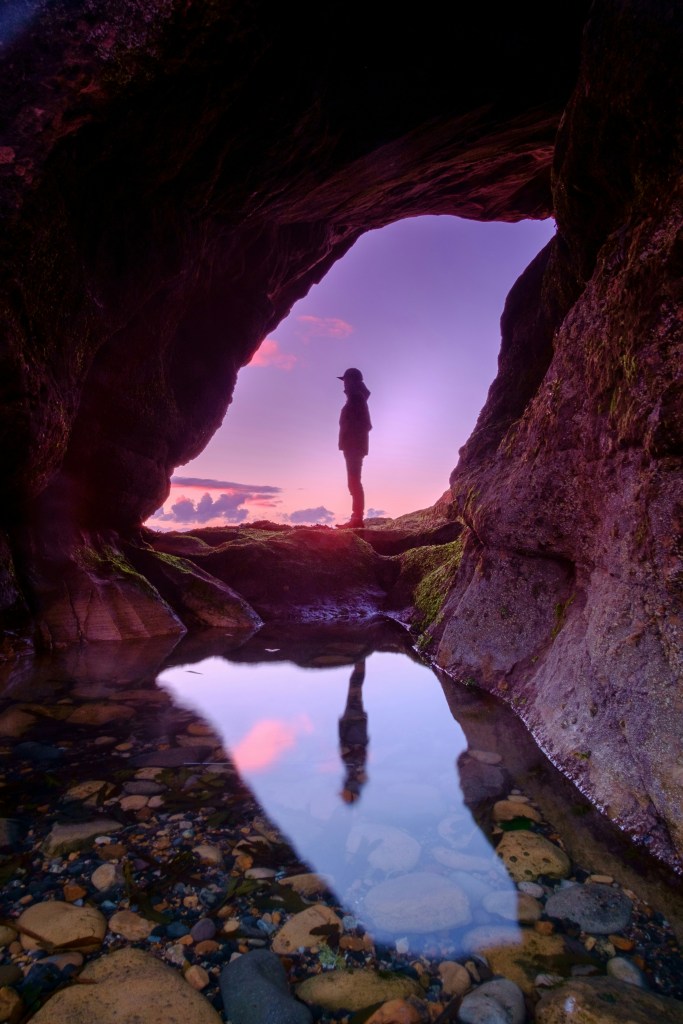 A person in hiking gear, is silhouetted by a purple sunset, they are framed by a circualr gap in the rockface as we look through a cave, and across a reflective pool, at them.