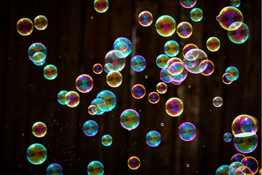 Photograph showing dozens of colourful, iridescent soap bubbles suspended against a dark background. Varied bubble sizes, overlapping clusters and rainbow reflections highlight delicate surfaces and floating motion.